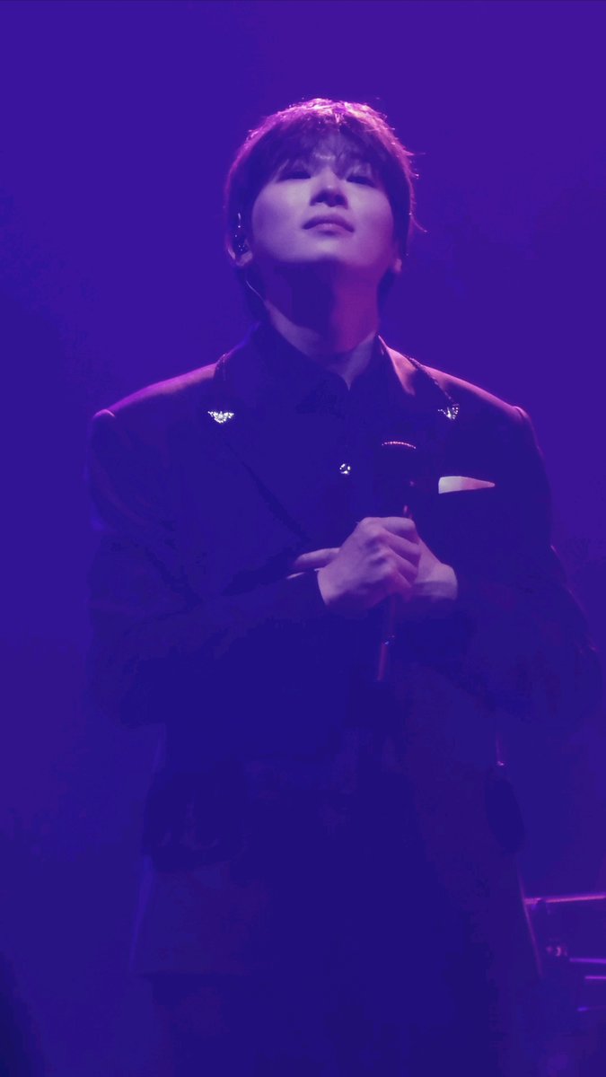A concert photo of an Asian male performer in a dark suit holding a microphone, illuminated by purple/violet stage lighting.