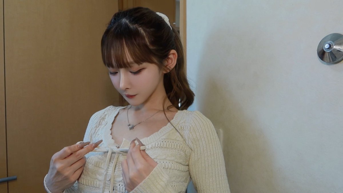 A young woman with brown hair tied back with a white scrunchie examines a small item, possibly an earring or piece of jewelry, while wearing a cream-colored knit cardigan with a delicate necklace, illustrating the post's appreciation for feminine experiences like accessorizing.