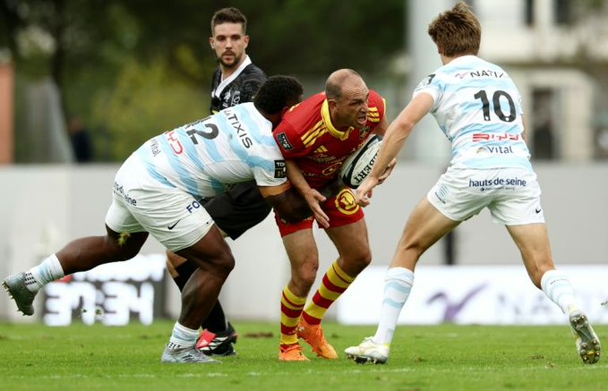 Action shot from a rugby match showing a player in a red and yellow jersey being tackled by two opposing players in light blue and white uniforms.