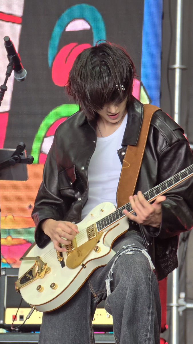 A guitarist with dark, shaggy hair performs on stage wearing a black leather jacket and white t-shirt, playing a cream-colored electric guitar with gold hardware.
