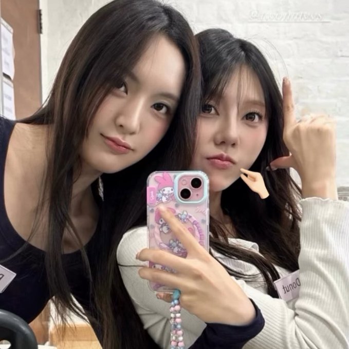 Two young women take a mirror selfie with a pink cartoon phone case, one wearing a black tank top and the other a white top with a 'twice' nametag.