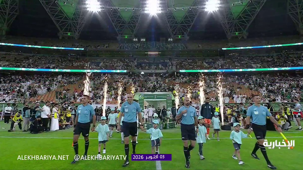 Four referees in light blue jerseys walk onto the field hand-in-hand with young mascots as cold pyrotechnic flames erupt in the background during the Saudi Arabia vs. Qatar match, with the Al-Ekhbariya Network watermark "ALEKHBARIYA.NET @ALEKHBARIYATV" and timestamp "3 أكتوبر 2026" visible at the bottom. The image captures the ceremonial entrance preceding the match referenced in the post, setting the stage for the first-half action between Al-Akhdar and Abu Nadi's squad.
