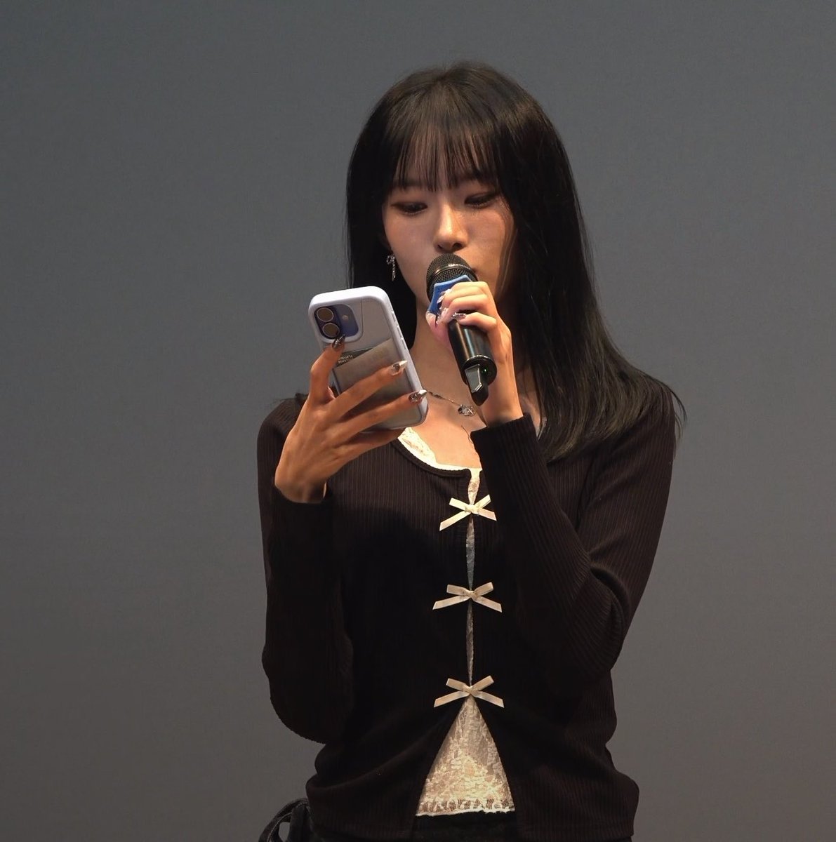 A person with long dark hair holding a microphone and a smartphone, wearing a black cardigan with white bow details over a lace top.