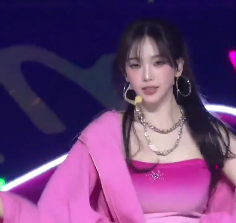 A young woman performs on stage wearing a bright pink dress, a pink jacket, large silver hoop earrings, and layered silver chain necklaces, with a headset microphone visible.