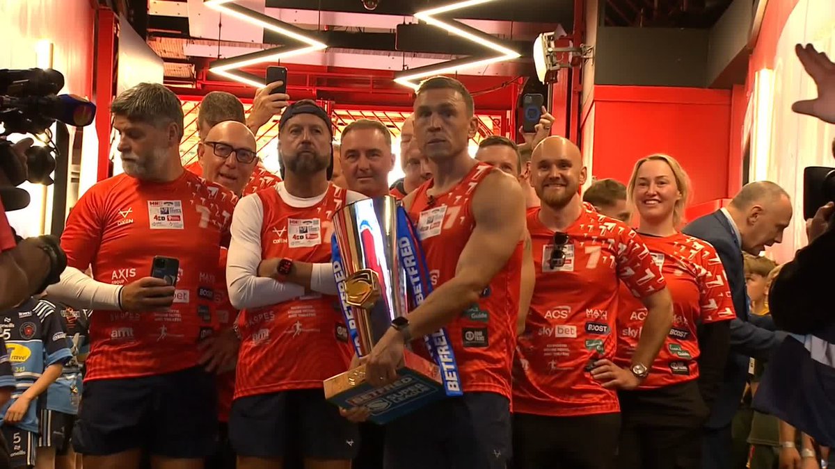 A muscular man in a red sports jersey proudly holds a Betfred Super League trophy while surrounded by teammates wearing red jerseys adorned with sponsors like ACS, Max, Sky Bet, and Boost, capturing a celebratory rugby league moment related to Sir Kevin Sinfield's charitable fundraising efforts.