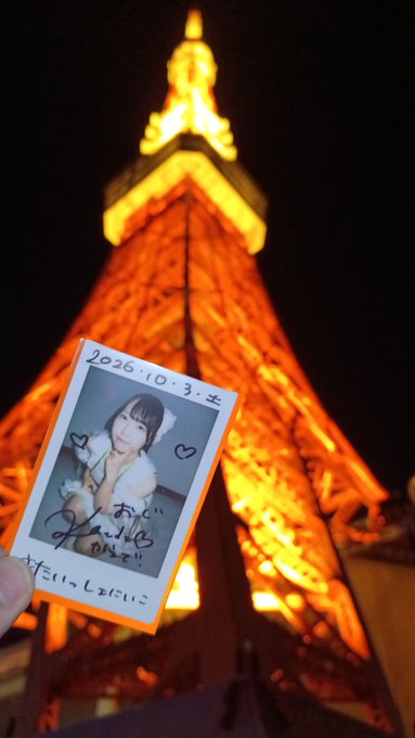 A hand holding a Polaroid photo of a dark-haired idol dated "2026.10.3" with Japanese writing and a heart, photographed in front of the illuminated Tokyo Tower at night.
