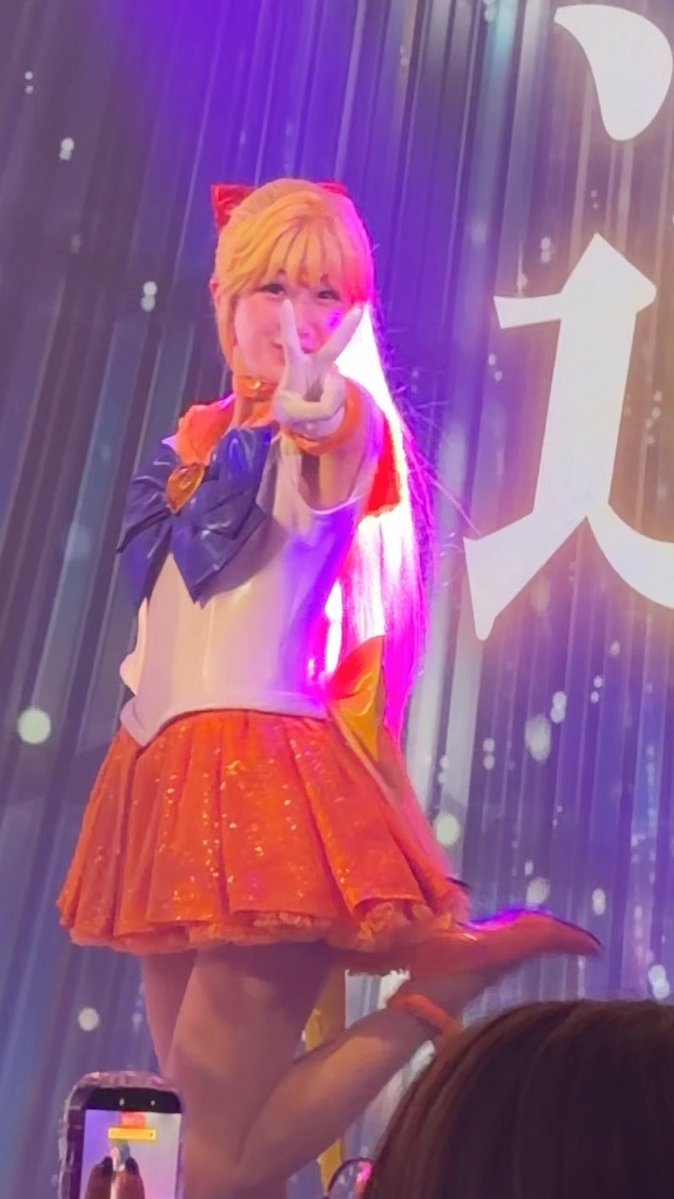 A performer dressed as Sailor Venus in an orange pleated skirt and sailor uniform posing on stage with a peace sign, against a purple backdrop with Japanese text.