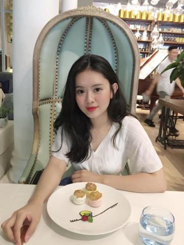 A young woman with long dark hair wearing a white top sits in an ornate light blue high-back chair at a restaurant table, where a plate of three small pastries and a glass of water are placed in front of her.