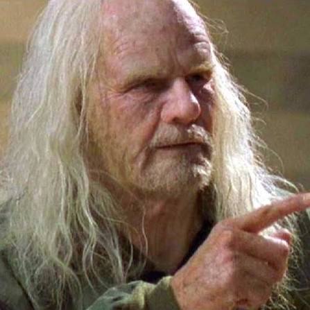 An older man with long white hair and beard pointing his finger, appearing to be from a film scene.