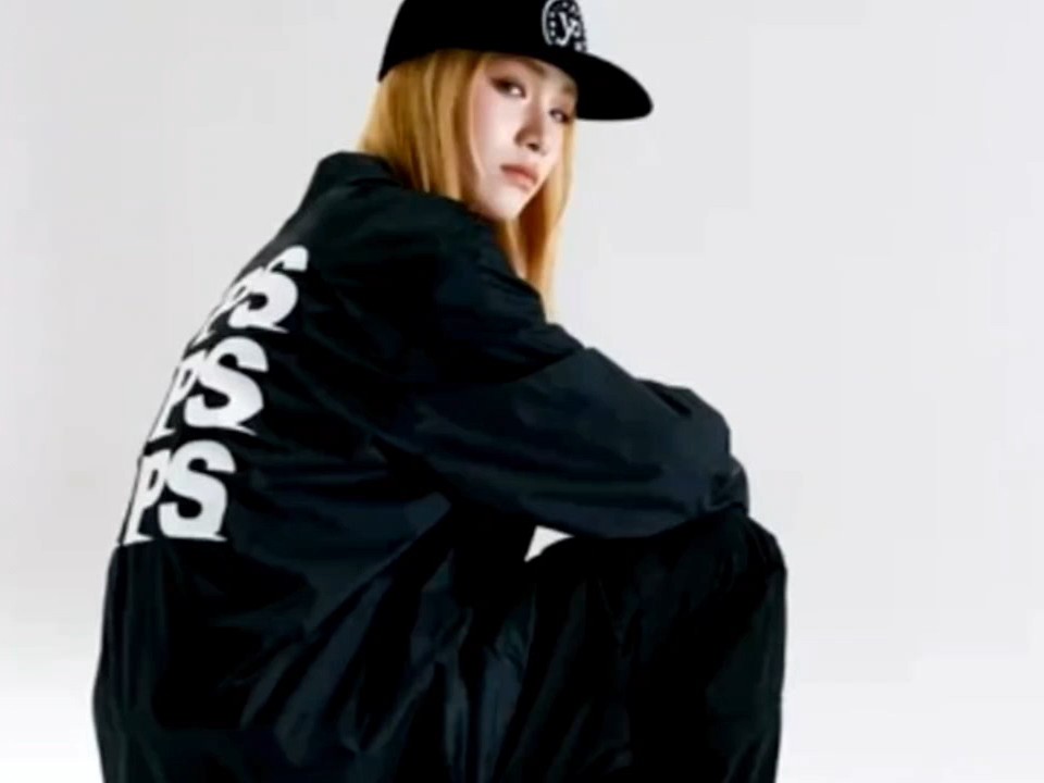 A blonde woman wearing a black cap and black jacket sits in a crouched pose against a white background, with 'BAPES' written vertically on her jacket.