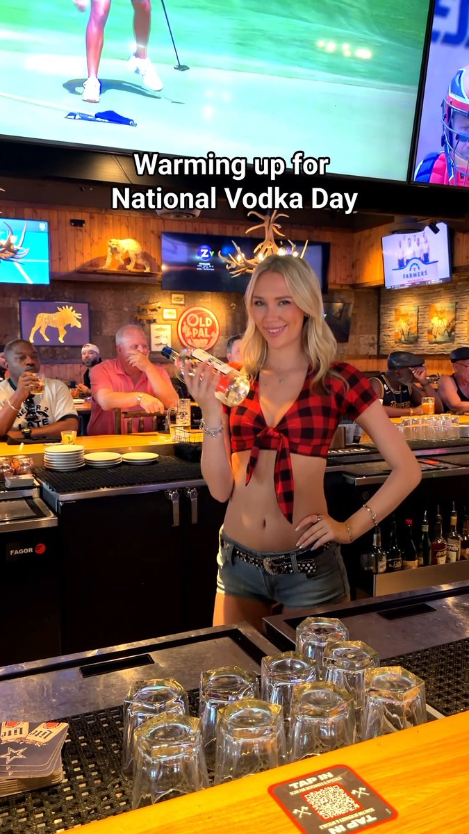 A blonde woman in a red plaid tied top and denim shorts holds a vodka bottle behind a bar, with text reading "Warming up for National Vodka Day" overlaid; TVs show sports and "OLD PAL" signage is visible.
