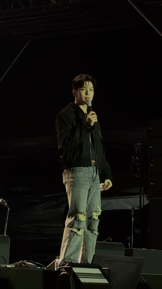 A man in a black jacket and ripped blue jeans holding a microphone while performing on a dark stage.