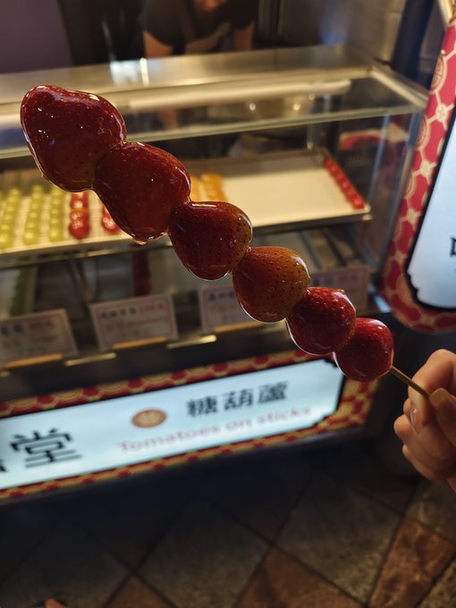 A hand holds a candied fruit skewer (tanghulu) in front of a shop display with a sign reading 'Tomato on sticks'.