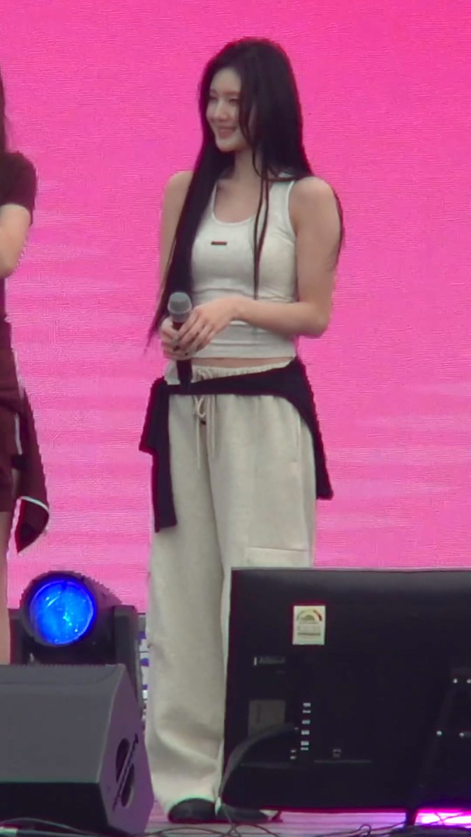 Young woman with long black hair standing on stage against a pink backdrop, wearing a white tank top and white sweatpants with a black sweater tied around her waist, holding a microphone.