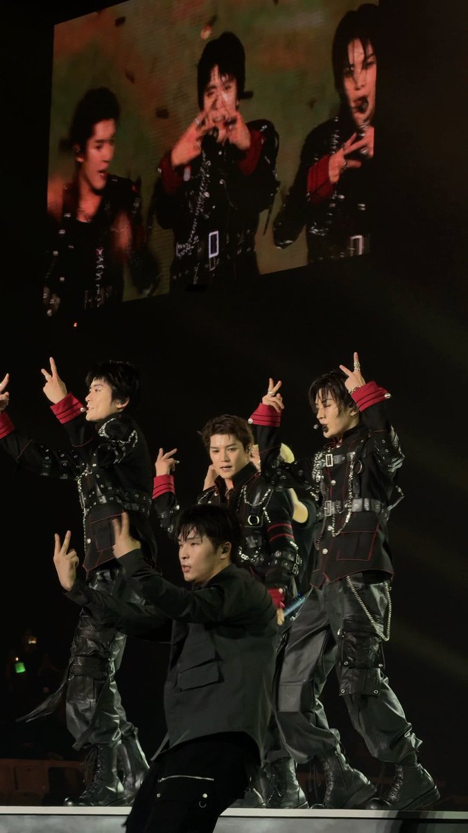 A group of performers in black outfits performing in front of a large screen displaying their images.