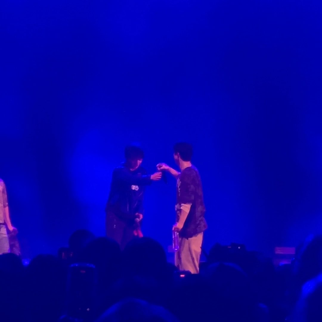 Two male performers interact on stage under deep blue lighting, with one handing a water bottle to the other in front of an audience.