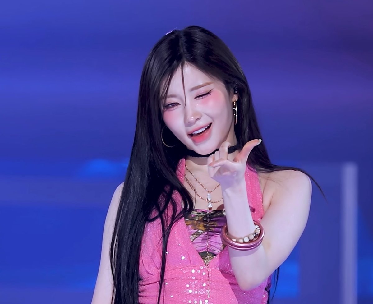 A close-up photo of a K-pop idol, likely from the group ILLIT, performing on stage with long dark hair, wearing a sparkly pink top, gold jewelry, and crying with a poignant expression while pointing.