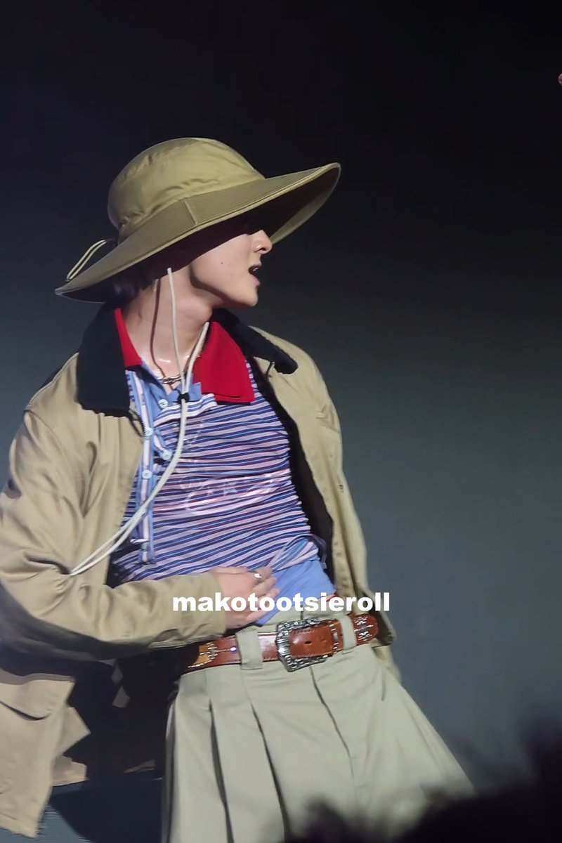 A concert photo of a performer wearing a wide-brimmed olive hat, a tan jacket, striped polo shirt, and belted pants, with a watermark reading 'makotootsieroll'.