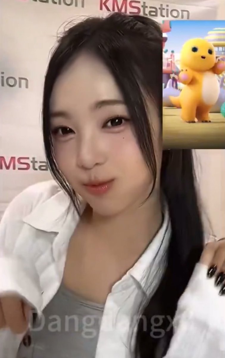 A selfie-style photo of a young woman with long dark hair wearing a white shirt, with a small inset image of a yellow cartoon character (from KMStation branding visible in the background) and watermark text 'Dang Longx'.