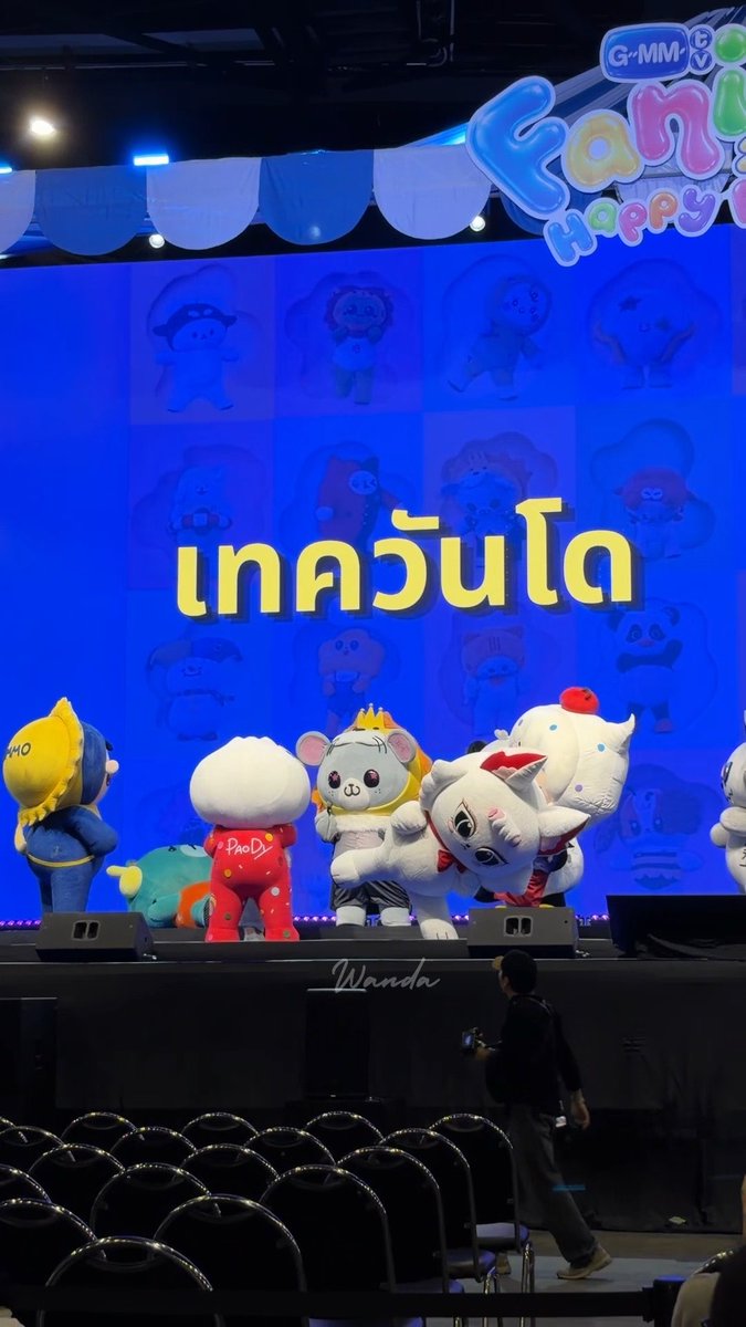 A lineup of colorful mascot characters performs on a stage with a vibrant blue backdrop featuring cartoon characters and the text "เทควันโด" (Taekwondo in Thai) alongside the "G-MM" logo, with an empty arena visible in the foreground where a photographer is capturing the show as part of the GMMTV FANIVAL 2026 Happy Family event.