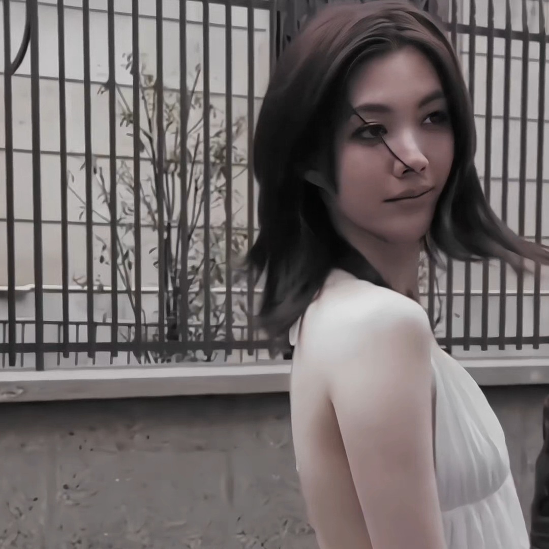 A portrait of a woman with long dark hair wearing a white sleeveless top, looking back over her shoulder against a backdrop of a window with metal grilles and foliage.