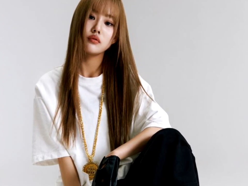 A young woman with straight light brown hair and bangs poses in a white t-shirt, wearing a long gold chain necklace with a pendant.