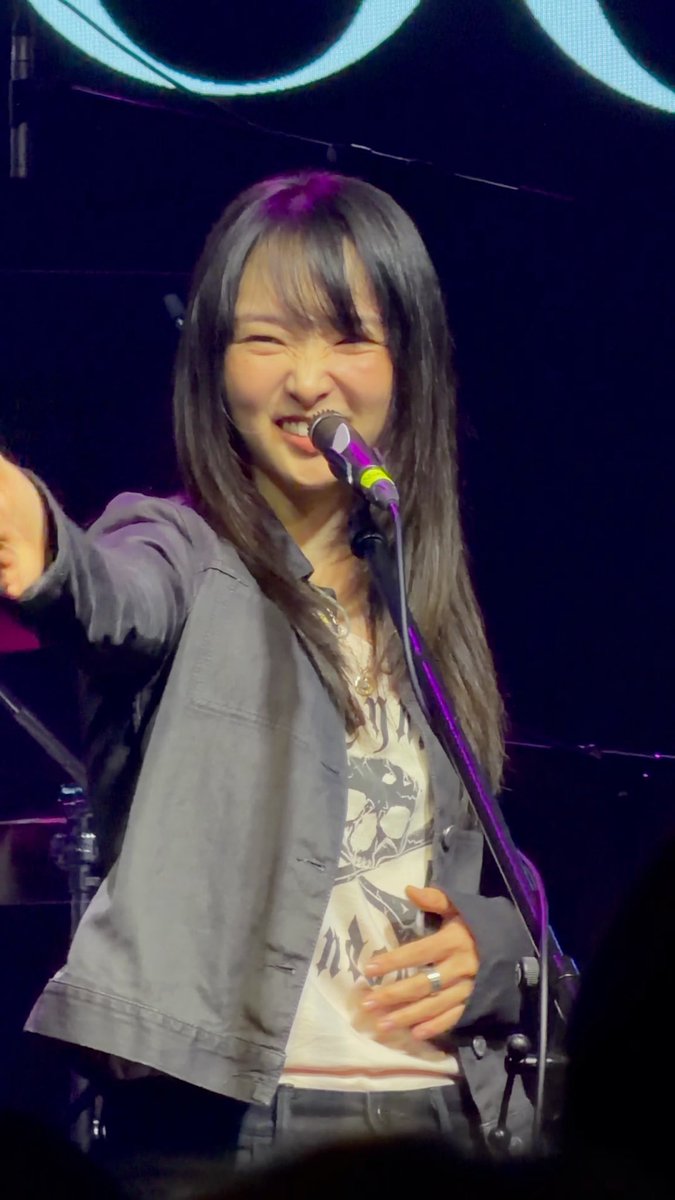 A young woman with long dark hair sings enthusiastically into a microphone on stage, wearing a denim jacket over a graphic t-shirt, with a ring visible on her finger.