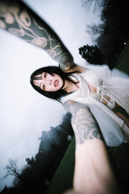 A photo taken from an unusual angle showing a smiling woman with dark hair and full arm tattoos wearing a white top, outdoors with trees in the background.