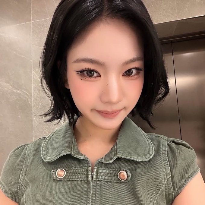 A close-up selfie of a young woman with short dark hair, dramatic eyeliner, and rosy cheeks wearing an olive green top with button details.