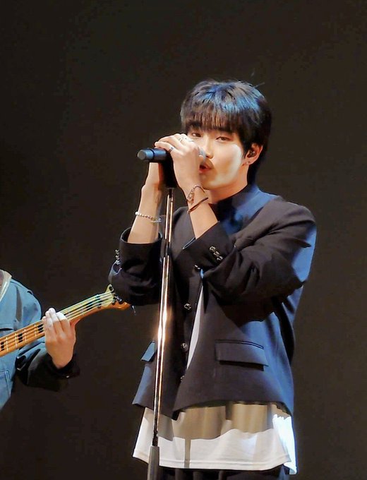 A young man in a dark blazer sings into a microphone on a stand, with another person partially visible playing a guitar to the left.