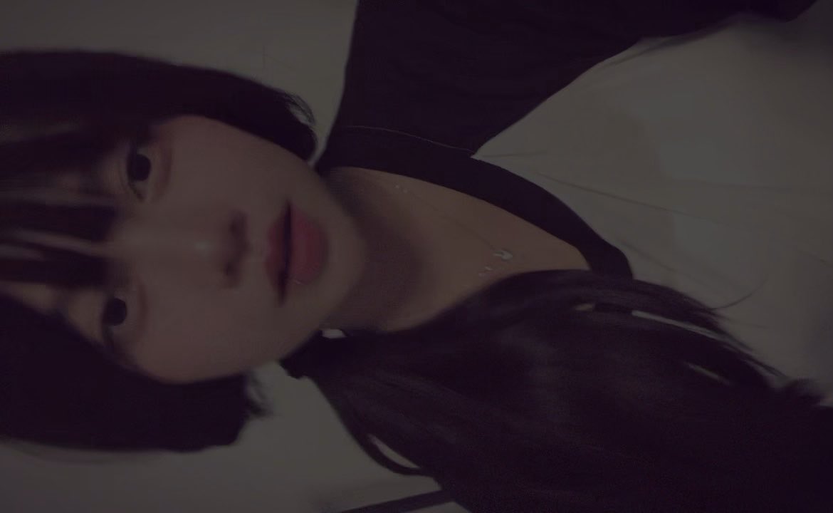 A dimly lit selfie of a young woman with long dark hair, taken at a rotated angle, wearing a black top and a delicate necklace.
