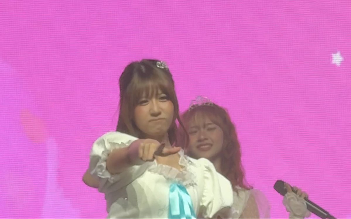 Two women in white outfits with crowns stand against a pink background; the woman in front makes a playful face and points at the camera while the other smiles behind her.