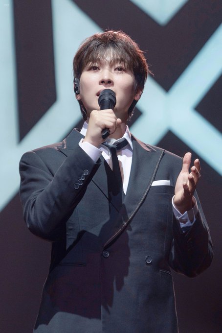 A male performer in a dark suit and tie singing into a microphone on stage with a geometric patterned backdrop.