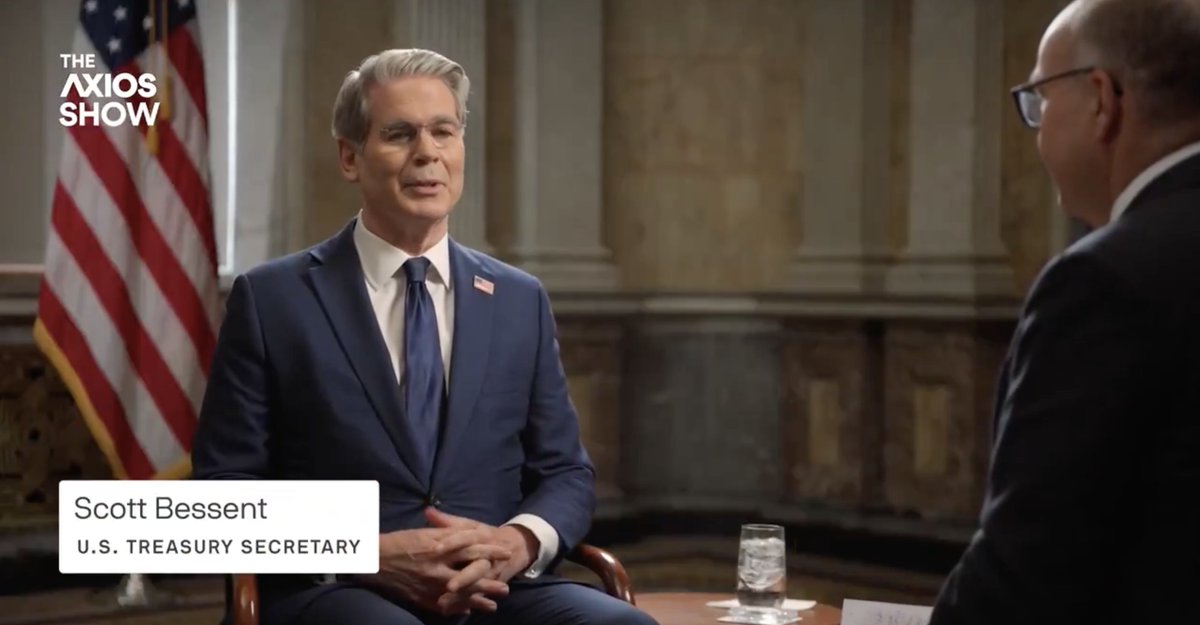 The image shows U.S. Treasury Secretary Scott Bessent being interviewed on The Axios Show, identified by the on-screen caption "Scott Bessent, U.S. Treasury Secretary" and the show's logo in the upper left corner. He is seated in a formal setting with an American flag in the background, wearing a navy blue suit and tie, while facing his interviewer across a small table with a water glass. The scene captures a moment from the extended interview referenced in the accompanying post.