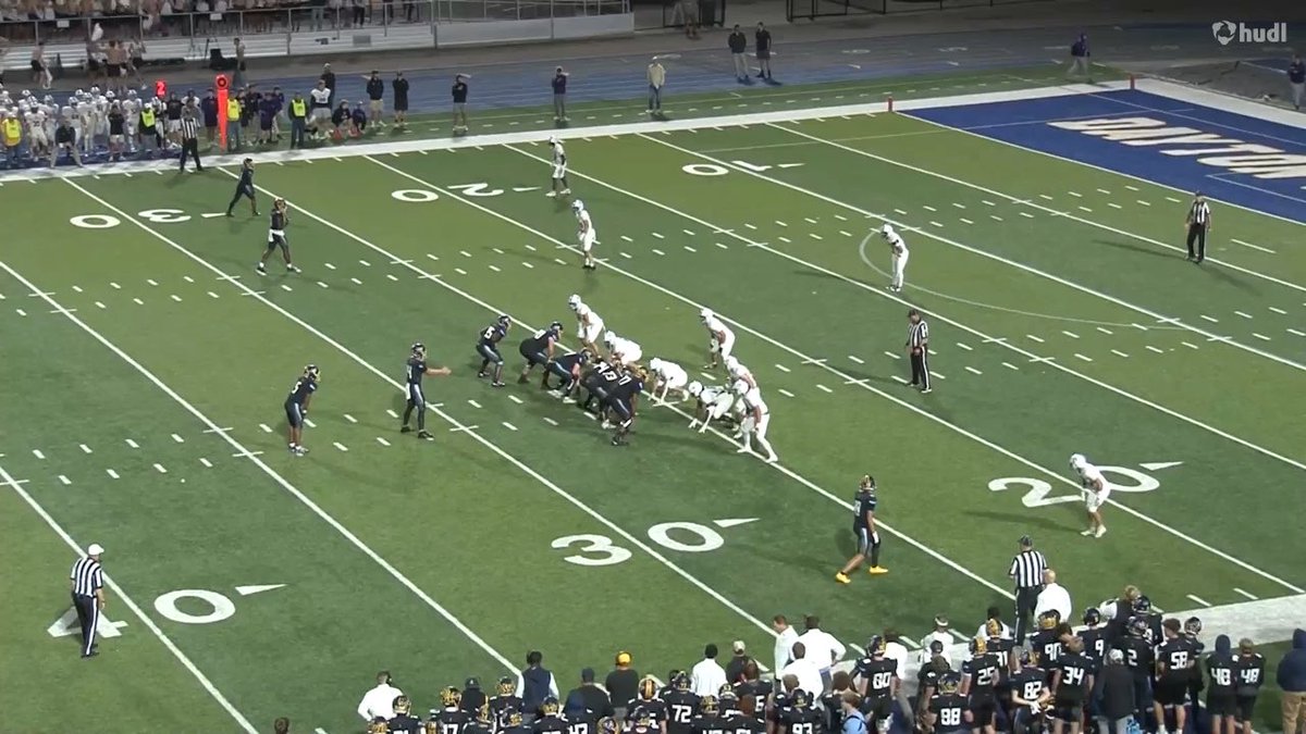 A high school football game is shown from an elevated sideline view, with the offensive team lined up near the 30-yard line while players, referees, and spectators watch from the sidelines.