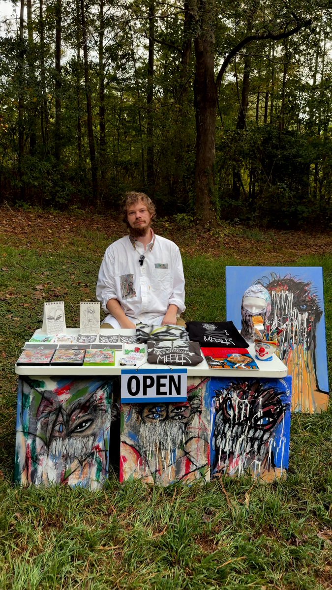An artist with curly hair and a beard sitting behind a table displaying his artwork, including paintings of crying faces, t-shirts, and a prominent 'OPEN' sign, set in a wooded outdoor area.