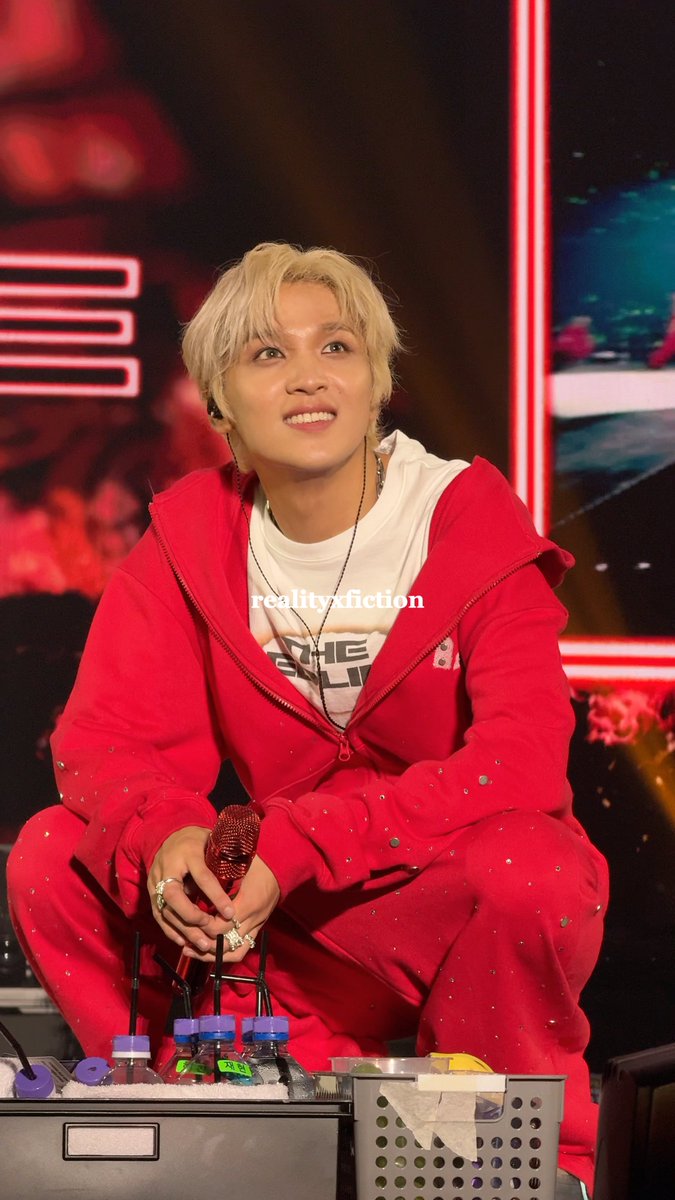 Haechan of NCT127 beams with a warm, loving expression during a stage appearance in Jakarta, wearing a striking red hooded jacket over a white t-shirt while holding a red microphone. The image captures his signature bright energy that fans adore, perfectly matching the affectionate "look of love" sentiment tagged with #NCT127inJAKARTA.