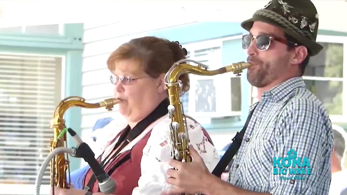 Two musicians play saxophones outdoors, with a 'KONA BIG WAVE' beer logo visible in the corner.