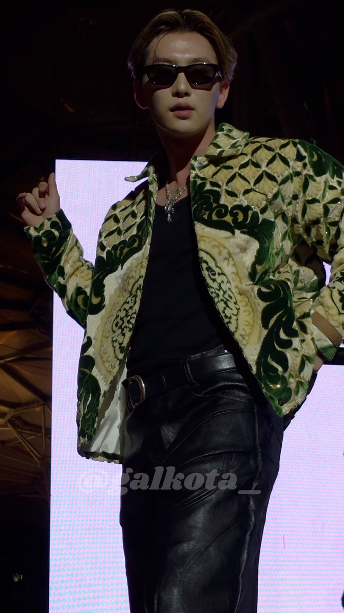 A performer wearing sunglasses, black leather pants, and an ornate green and gold patterned jacket poses on stage with 'galkota_' watermark visible.