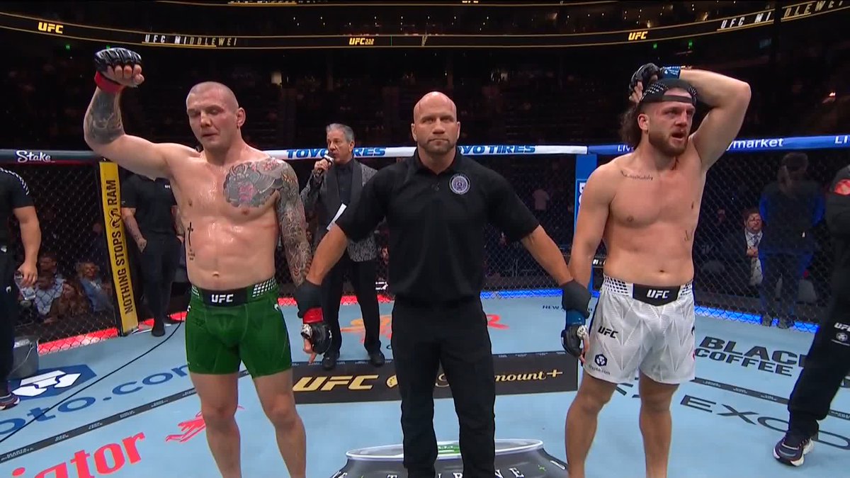 A referee raises the arms of two shirtless UFC fighters inside the octagon after their bout, with the fighter on the left wearing green shorts and the fighter on the right wearing white shorts, while "UFC," "Stake," and "Black Coffee" branding is visible on the cage and mat. The image captures the official decision announcement moment, likely showing Ismail Naurdiev (left) being declared the winner over Marvin Vettori (right) by unanimous decision at UFC 332.