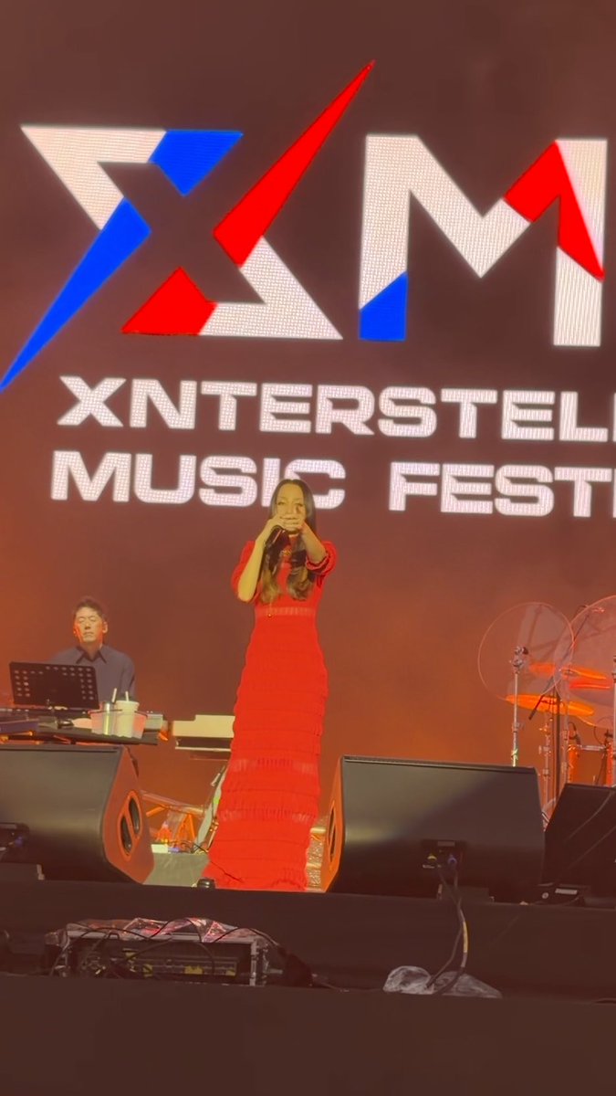 A female singer in a long red dress performs on stage at the 'XM' Xnterstellar Music Festival, with a band and drum kit visible behind her.