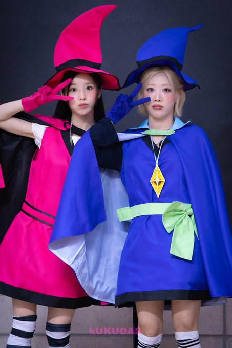 Two women pose in witch cosplay costumes, one in a pink and black outfit and the other in a blue outfit with a green bow and yellow pendant, both making peace signs, with the watermark 'KUKUDA' visible at the bottom.