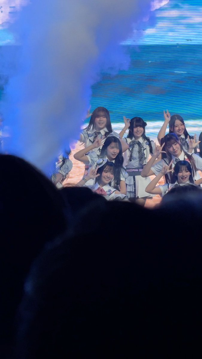 A group of young women in school uniforms pose with peace signs on a stage with blue lighting, viewed from the audience.