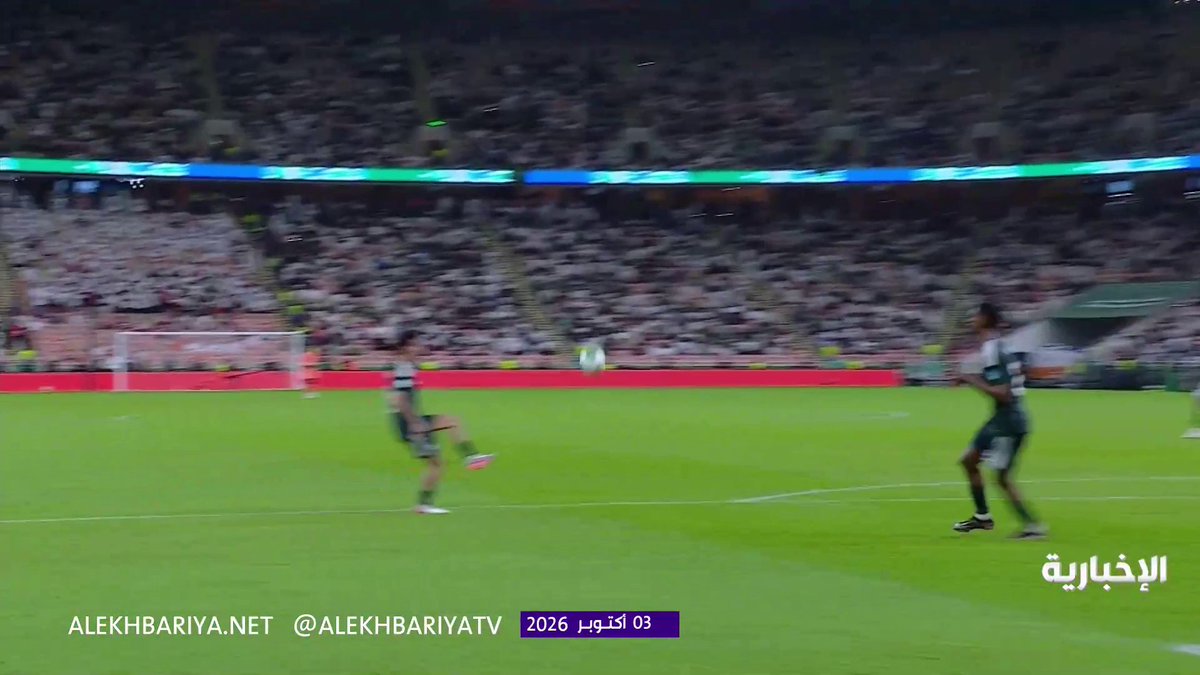 A soccer player in green and white kit kicks the ball in front of a crowded stadium, while the watermark "ALEKHBARIYA.NET @ALEKHBARIYATV" and date "03 اكتوبر 2026" appear at the bottom, contextualizing the moment as part of the Saudi Arabia vs. Qatar match where Al-Akhdar's players were appealing for a penalty that the referee denied.