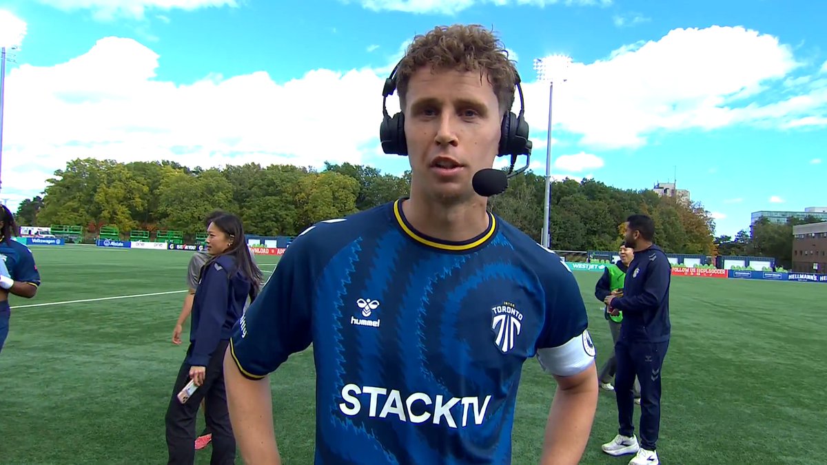 A curly-haired soccer player wearing headphones and a microphone conducts a postgame interview on the field, wearing a blue "INTER TORONTO" jersey with "STACKTV" and "hummel" branding visible. The image aligns with the caption about Frank Sturing's interview following Inter Toronto's 3-1 victory over Cavalry in the Canadian Premier League.