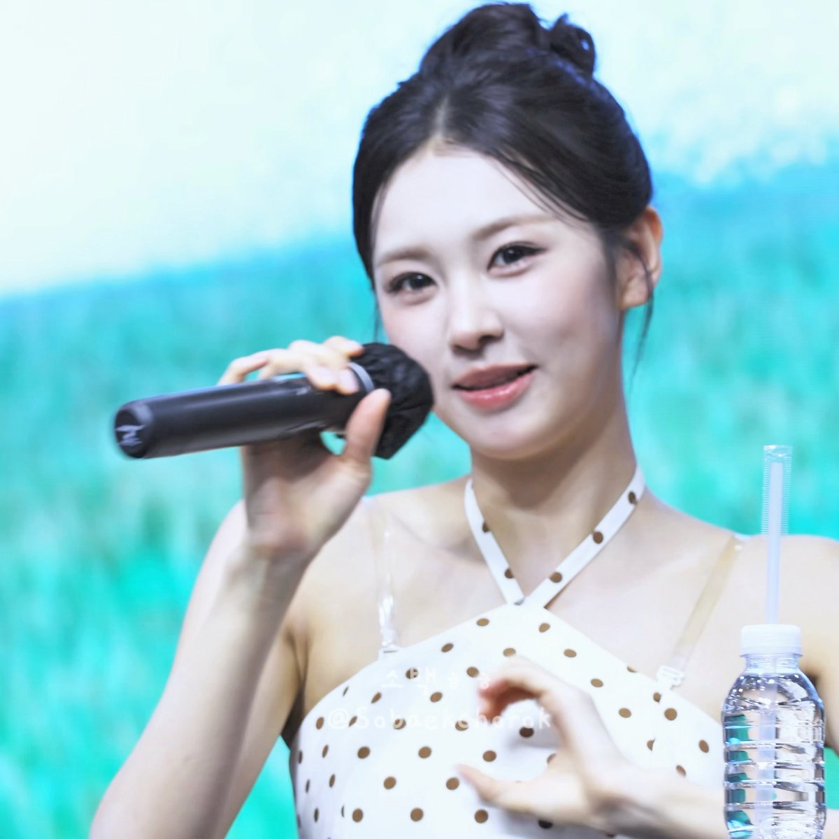 A woman with dark hair in a bun holds a microphone while speaking, wearing a white dress with brown polka dots, with a water bottle visible nearby.