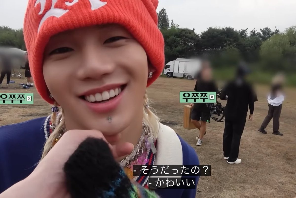 A video still image of a smiling woman wearing an orange beanie and chunky chain necklace at an outdoor festival, with Japanese subtitles reading '- そうだったの？' and '- かわいい'.