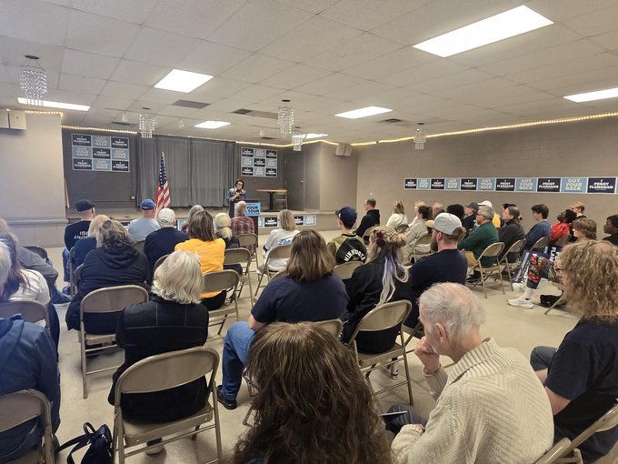 A speaker addresses a packed room of supporters at what appears to be a campaign event for Minnesota Lt. Governor Peggy Flanagan, part of her "Got Your Back" tour just one month before Election Day. Signs visible in the crowd read "Flanagan" alongside the slogan "got your back," reflecting her grassroots campaign that has engaged over 6,000 volunteers across 112+ Minnesota communities.