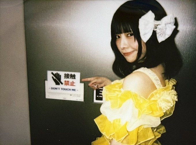 A young woman with dark hair wearing a white bow and a yellow ruffled dress, pointing at a 'DON'T TOUCH ME' sign with Japanese text on a dark door.
