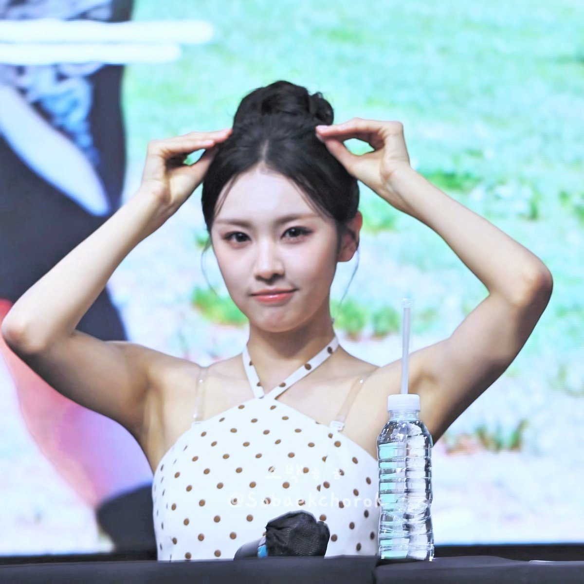 A young woman with her hair in a bun poses with her hands raised to her head at what appears to be a fan event, wearing a white polka-dot halter top, with a water bottle on the table and a watermark '@Sobaekchorok'.